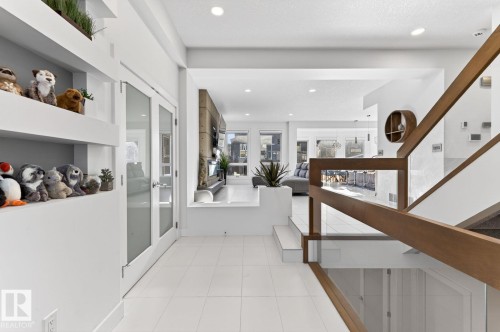Contemporary open-concept interior featuring a glass and wood-finish staircase, white tile flooring, built-in shelving, recessed lighting, and a double-door entry with frosted glass - 793 Windermere Wynd, Edmonton, AB - Indoor Photo Showing Other Room