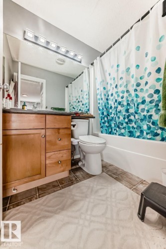 Bathroom featuring a light wood-finish vanity with a dark countertop, an integrated mirror with a six-light fixture, a white toilet, and a white bathtub with an overhead shower - 715 Hudson Place, Edmonton, AB - Indoor Photo Showing Bathroom