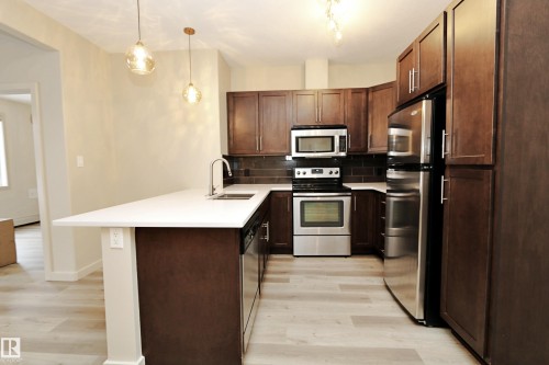 Contemporary kitchen featuring dark wood cabinetry, stainless steel appliances, light-toned wood-finish flooring, a white countertop, and a dark subway tile backsplash - 140 308 Ambleside Link Link Sw, Edmonton, AB - Indoor Photo Showing Kitchen With Double Sink