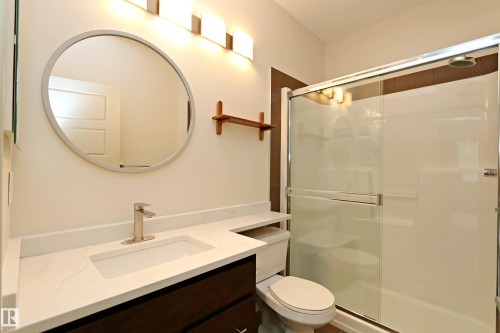Contemporary bathroom featuring a dark wood-finish vanity with a white countertop and integrated sink, brushed nickel faucet, and a large circular mirror - 140 308 Ambleside Link Link Sw, Edmonton, AB - Indoor Photo Showing Bathroom