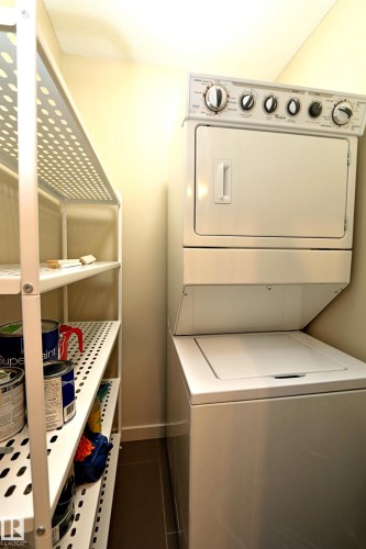 Compact laundry area featuring a stacked washer and dryer unit, complementary shelving, and dark tile flooring - 140 308 Ambleside Link Link Sw, Edmonton, AB - Indoor Photo Showing Laundry Room