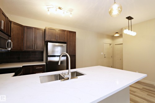 Kitchen featuring a large central island with a double stainless steel sink and a gooseneck faucet, dark wood-finish cabinetry, stainless steel appliances, and a dark tile backsplash - 140 308 Ambleside Link Link Sw, Edmonton, AB - Indoor Photo Showing Kitchen With Double Sink