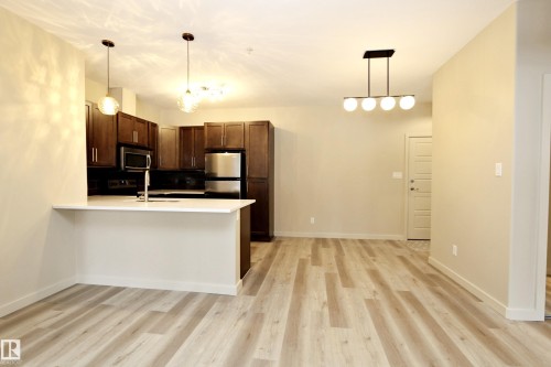 Open-concept living space featuring wood-finish flooring, a kitchen island with a sink, dark wood cabinetry, stainless steel appliances, and contemporary lighting fixtures - 140 308 Ambleside Link Link Sw, Edmonton, AB - Indoor Photo Showing Kitchen