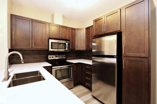 Contemporary kitchen featuring dark wood cabinetry, stainless steel appliances, white countertops, and a subway tile backsplash - 140 308 Ambleside Link Link Sw, Edmonton, AB - Indoor Photo Showing Kitchen With Double Sink