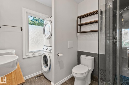 Modern bathroom featuring a stacked laundry unit, a glass-enclosed shower with dark subway tile, a white toilet, and wood-finish wall shelving - 13635 108 Avenue, Edmonton, AB - Indoor Photo Showing Laundry Room