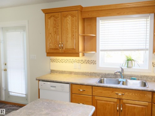 Kitchen featuring natural wood cabinetry with crown molding, double basin stainless steel sink, built-in dishwasher, and a window with blinds - 5802 52 Avenue, Barrhead, AB - Indoor Photo Showing Kitchen With Double Sink