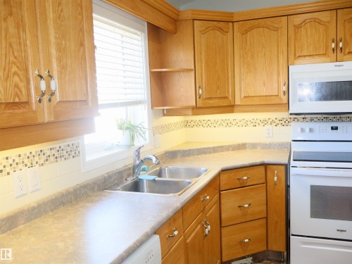 Kitchen workspace featuring wood-finish cabinetry, a double-basin stainless steel sink with a gooseneck faucet, and a mosaic tile backsplash - 5802 52 Avenue, Barrhead, AB - Indoor Photo Showing Kitchen With Double Sink