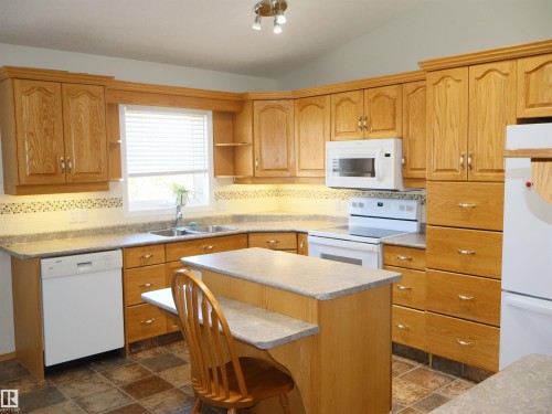 Kitchen featuring extensive wood cabinetry, a central island with a breakfast bar, white appliances, and tiled flooring - 5802 52 Avenue, Barrhead, AB - Indoor Photo Showing Kitchen With Double Sink