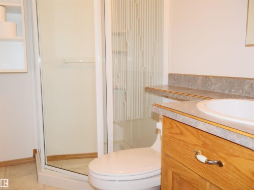 Bathroom featuring a walk-in shower with clear glass enclosure and textured privacy panel, a toilet, a vanity with a white basin and gray-patterned countertop, and wood-finish cabinetry - 5802 52 Avenue, Barrhead, AB - Indoor Photo Showing Bathroom