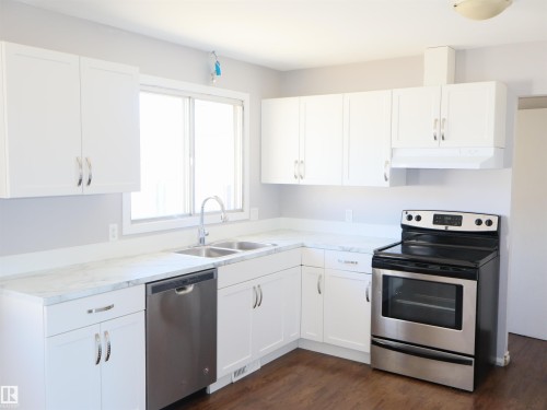Kitchen featuring white shaker-style cabinetry, a double basin stainless steel sink, stainless steel dishwasher, stainless steel range, and wood-finish flooring - 4934 49 Avenue, Barrhead, AB - Indoor Photo Showing Kitchen With Double Sink