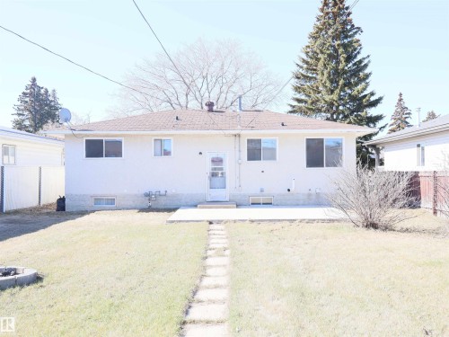Rear exterior featuring a white stucco finish, brown shingle roof, multiple windows, and a back door leading to a concrete patio - 4934 49 Avenue, Barrhead, AB - Outdoor