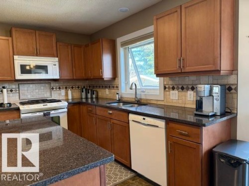 Kitchen featuring rich wood cabinetry, dark speckled countertops, and a tiled backsplash with decorative accents - 356 Weber Way, Edmonton, AB - Indoor Photo Showing Kitchen With Double Sink