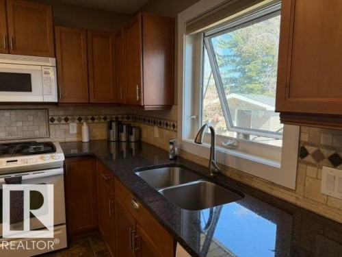 Kitchen featuring a double basin stainless steel sink with a gooseneck faucet, dark speckled countertops, rich wood-finish cabinetry, and a mosaic tile backsplash - 356 Weber Way, Edmonton, AB - Indoor Photo Showing Kitchen With Double Sink
