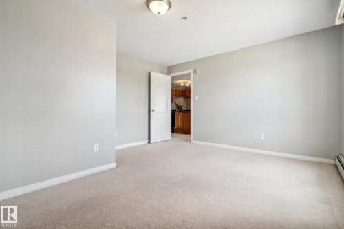 Carpeted room featuring light gray wall paint, white trim, a semi-flush ceiling light fixture, and a doorway opening to a kitchen with wood-finish cabinetry - 1306 330 Clareview Station Drive, Edmonton, AB - Indoor Photo Showing Other Room