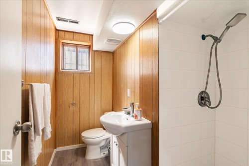 Bathroom featuring wood-panel walls, a window with security bars, a white pedestal sink, a toilet, and a shower with white tiled walls and a handheld showerhead - 10531 128 Avenue, Edmonton, AB - Indoor Photo Showing Bathroom