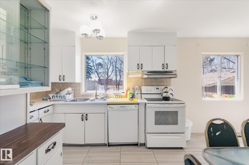 Well-lit kitchen featuring white cabinetry, a built-in dishwasher, white electric range, and a dual-basin sink with a window overhead - 10531 128 Avenue, Edmonton, AB - Indoor Photo Showing Kitchen With Double Sink