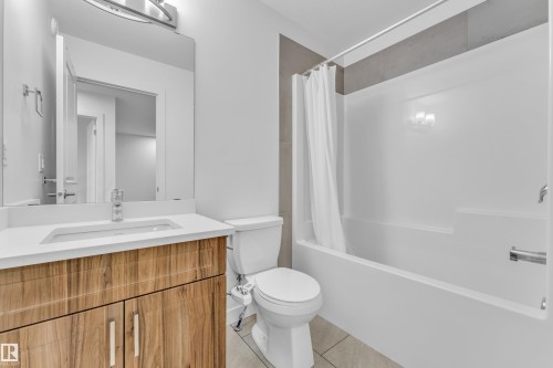 Contemporary bathroom featuring a wood-finish vanity with a white countertop and an integrated sink, a large wall-mounted mirror, and a white bathtub with a shower and gray tiled surround - 41 603 Orchards Boulevard, Edmonton, AB - Indoor Photo Showing Bathroom