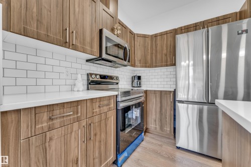Kitchen featuring extensive wood-finish cabinetry, white subway tile backsplash, white countertops, stainless steel appliances, and wood-finish flooring - 41 603 Orchards Boulevard, Edmonton, AB - Indoor Photo Showing Kitchen