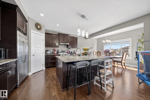 Open-concept kitchen featuring an island with pendant lighting and seating, rich wood-finish flooring, dark wood cabinetry, stainless steel appliances, and a dining area with chandelier lighting and a large window - 3639 15A Street, Edmonton, AB - Indoor Photo Showing Other Room