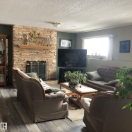 Living area featuring a floor-to-ceiling stacked stone fireplace with a wood mantle and brass-toned screen, wood-finish flooring, recessed window, and wall-mounted lighting - 
