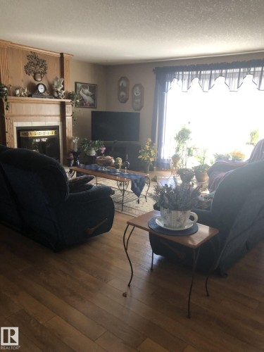 Living room featuring wood-finish flooring, a fireplace with a stone surround and wood mantle, and a large window with valance - 123064 Hwy 45, Rural Two Hills County, AB 