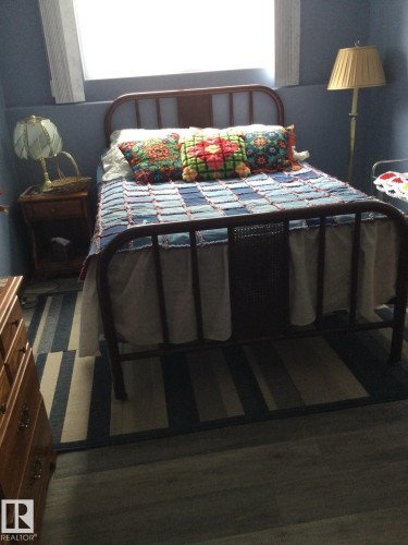 Wood-finish flooring with a striped area rug, complemented by a metal frame bed and a window with white blinds - 123064 Hwy 45, Rural Two Hills County, AB 