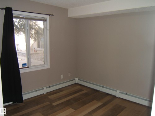 Wood-finish flooring throughout, featuring a window with white trim, baseboard heating, and neutral wall tones - 105 11214 80 Street, Edmonton, AB - Indoor Photo Showing Other Room