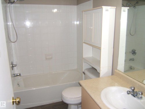Bathroom featuring a built-in tub with tile surround, a single vanity with a laminate countertop, and wood-finish flooring - 105 11214 80 Street, Edmonton, AB - Indoor Photo Showing Bathroom