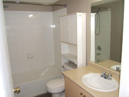 Bathroom featuring a white tiled bath and shower combination, a white commode, and a light-toned vanity with an integrated sink and chrome faucet - 105 11214 80 Street, Edmonton, AB - Indoor Photo Showing Bathroom