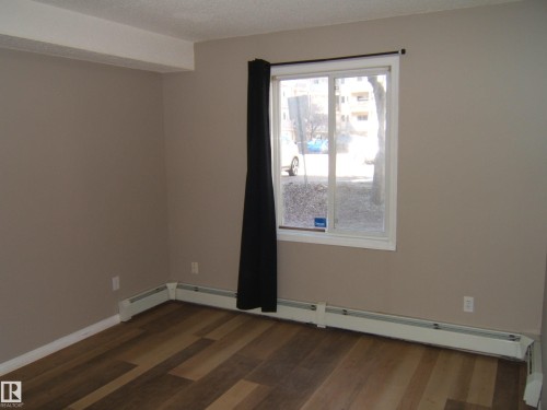 Room featuring wood-finish flooring, a large window, neutral wall paint, white trim, and baseboard heating - 105 11214 80 Street, Edmonton, AB - Indoor Photo Showing Other Room