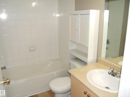 Bathroom with a white tiled tub surround, a built-in white over-toilet cabinet, and a vanity with an integrated sink and mirror - 105 11214 80 Street, Edmonton, AB - Indoor Photo Showing Bathroom