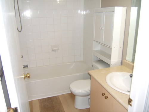 Bathroom featuring a white tiled tub/shower combination, over-toilet storage cabinetry, a single-sink vanity with a light-toned countertop, and wood-finish flooring - 105 11214 80 Street, Edmonton, AB - Indoor Photo Showing Bathroom