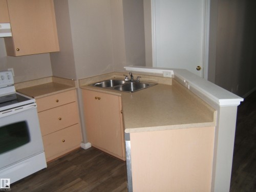 Kitchen featuring a double basin stainless steel sink, light-toned countertops, wood-finish flooring, and light peach cabinetry with brass hardware - 105 11214 80 Street, Edmonton, AB - Indoor Photo Showing Kitchen With Double Sink