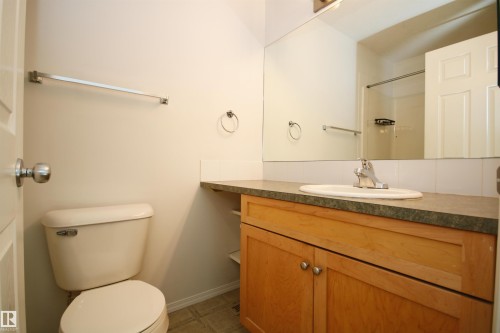 Bathroom featuring a natural wood-finish vanity with a dark countertop, an integrated sink, a large wall-mounted mirror, and a white toilet - 11 230 Edwards Drive, Edmonton, AB - Indoor Photo Showing Bathroom