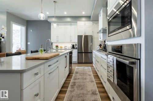 Spacious kitchen featuring an expansive center island with an undermount sink, white shaker cabinetry, stainless steel appliances, and wood-finish flooring - 108 7463 May Common, Edmonton, AB - Indoor Photo Showing Kitchen With Upgraded Kitchen