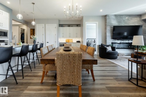 Open-concept interior featuring wood-finish flooring, a kitchen island with pendant lighting, white cabinetry, and a built-in electric fireplace with a stone surround - 108 7463 May Common, Edmonton, AB - Indoor Photo Showing Dining Room