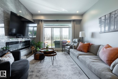 Living area featuring an accent wall with a mounted television above an electric fireplace, recessed lighting, and sliding glass doors opening to the exterior - 108 7463 May Common, Edmonton, AB - Indoor Photo Showing Living Room
