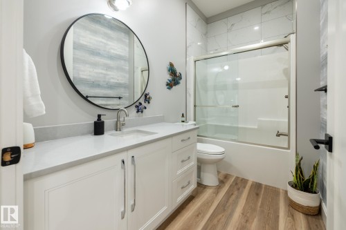 Bathroom featuring a white vanity with an undermount sink, a large round mirror, a full bathtub with a glass enclosure, a white toilet, and wood-finish flooring - 108 7463 May Common, Edmonton, AB - Indoor Photo Showing Bathroom