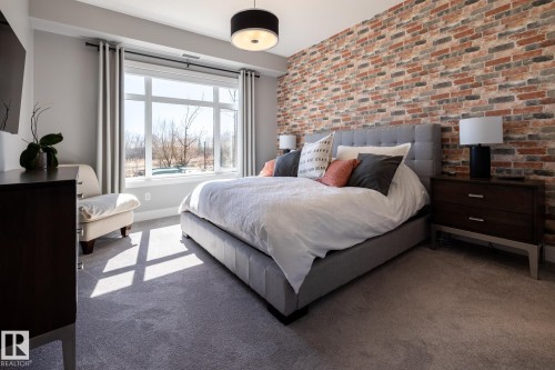 Bedroom featuring a multi-tone brick accent wall, large window with transom, grey carpet flooring, crown molding, and a contemporary ceiling light fixture - 108 7463 May Common, Edmonton, AB - Indoor Photo Showing Bedroom