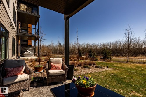 Ground-level patio featuring a covered outdoor area, direct lawn access, and natural landscaping beyond a black metal fence - 108 7463 May Common, Edmonton, AB - Outdoor