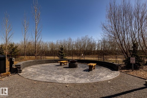 Outdoor patio featuring a circular paver surface, a curved retaining wall, a central fire pit, two wooden benches, and surrounding gravel landscaping - 108 7463 May Common, Edmonton, AB - Outdoor