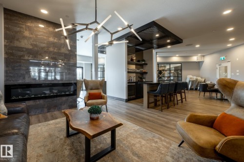 Spacious room featuring a contemporary linear fireplace set into a large-format tile accent wall, wood-finish flooring, a built-in wet bar with shelving, and a decorative ceiling treatment - 108 7463 May Common, Edmonton, AB - Indoor Photo Showing Living Room