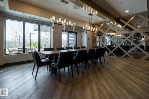 Spacious gathering area featuring a long wooden table with seating for twelve, wood-finish flooring, and a contemporary linear chandelier with multiple glass globes - 108 7463 May Common, Edmonton, AB - Indoor Photo Showing Dining Room