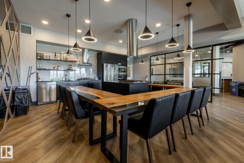 Spacious common area featuring wood-finish flooring, a contemporary kitchen with stainless steel appliances, and a large U-shaped live-edge wood table - 108 7463 May Common, Edmonton, AB - Indoor