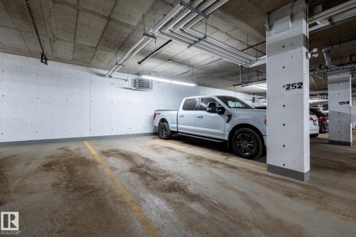 Underground parking space featuring concrete walls and ceiling, exposed utility piping, and fluorescent lighting - 108 7463 May Common, Edmonton, AB - Indoor Photo Showing Garage