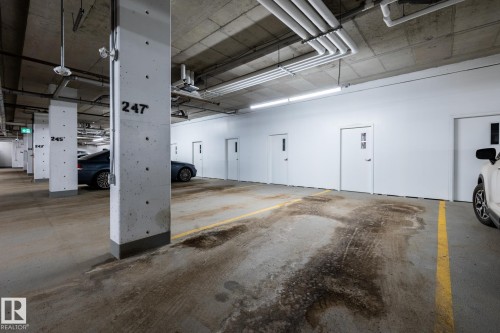Underground parking garage featuring concrete flooring, exposed ceiling utilities, bright fluorescent lighting, and white wall panels with multiple access doors - 108 7463 May Common, Edmonton, AB - Indoor Photo Showing Garage