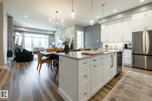 Open-concept living space featuring a gourmet kitchen with white shaker cabinetry, stainless steel appliances, and a central island with a light-toned countertop - 108 7463 May Common, Edmonton, AB - Indoor Photo Showing Kitchen With Upgraded Kitchen