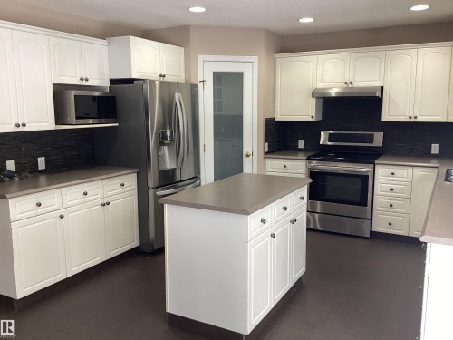 Modern kitchen featuring white shaker-style cabinetry, a central island, stainless steel appliances, dark stone-look countertops, and a dark tile backsplash - 54 Leonard Drive, St. Albert, AB - Indoor Photo Showing Kitchen