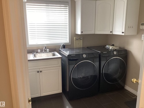 Utility room featuring a window with horizontal blinds, a stainless steel sink with a two-handle faucet, white cabinetry, dark countertop, and dark floor tiling - 54 Leonard Drive, St. Albert, AB - Indoor Photo Showing Laundry Room