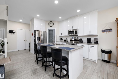 Contemporary kitchen featuring an island with seating, glossy white cabinetry, stainless steel appliances, a light-toned countertop, and wood-finish flooring - 82 Wingate Way, Fort Saskatchewan, AB - Indoor Photo Showing Kitchen